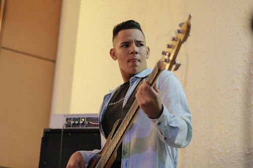 A man is playing an electric bass guitar while looking focused. He is wearing a light denim jacket over a black t-shirt. In the background, there is an amplifier, and the setting appears to be indoors, with textured cream-colored walls.