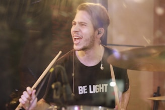 A man with short hair is passionately playing the drums. He is wearing a black T-shirt with the words 'BE LIGHT' printed in white. Earphones are visible in his ears as he holds a pair of drumsticks. His expression is intense, and there is a blur around him that suggests motion. The lighting creates a warm atmosphere.