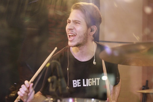 A man with short hair is passionately playing the drums. He is wearing a black T-shirt with the words 'BE LIGHT' printed in white. Earphones are visible in his ears as he holds a pair of drumsticks. His expression is intense, and there is a blur around him that suggests motion. The lighting creates a warm atmosphere.