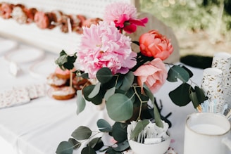 A beautifully arranged bouquet of vibrant pink and orange flowers with green leafy foliage is placed on a white table. The table also has stacked paper cups with gold patterns, a bowl filled with various stirrers or packets, and a background showcasing a pegboard with an assortment of donuts on display.