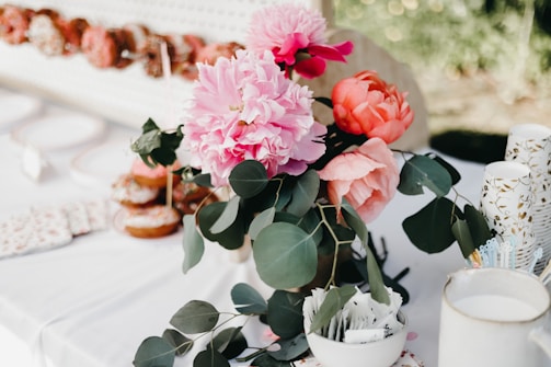 A beautifully arranged bouquet of vibrant pink and orange flowers with green leafy foliage is placed on a white table. The table also has stacked paper cups with gold patterns, a bowl filled with various stirrers or packets, and a background showcasing a pegboard with an assortment of donuts on display.
