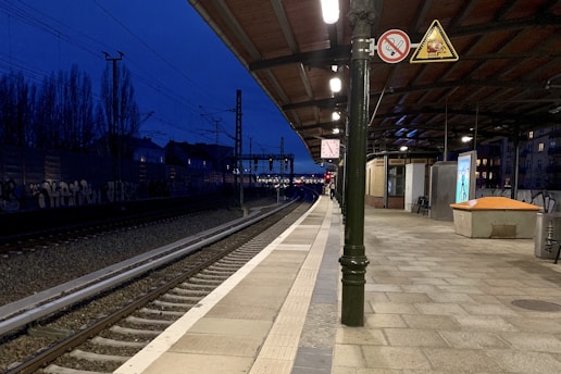 A railway platform during the evening with a deep blue sky. The platform is deserted, featuring a clock, metal railing, signage, and a shelter area for passengers. Electric tracks extend into the distance along graffiti-covered walls. Artificial lights illuminate the platform and tracks, providing contrast against the darkening sky.