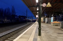 A railway platform during the evening with a deep blue sky. The platform is deserted, featuring a clock, metal railing, signage, and a shelter area for passengers. Electric tracks extend into the distance along graffiti-covered walls. Artificial lights illuminate the platform and tracks, providing contrast against the darkening sky.