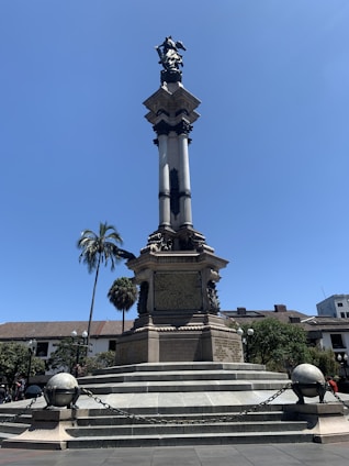 A tall stone monument with a statue on top, surrounded by a circular stone base and steps. The structure is adorned with inscriptions and framed by decorative chains and spheres. In the background, there is a clear blue sky, a palm tree, and several buildings, including one with a traditional architectural style.