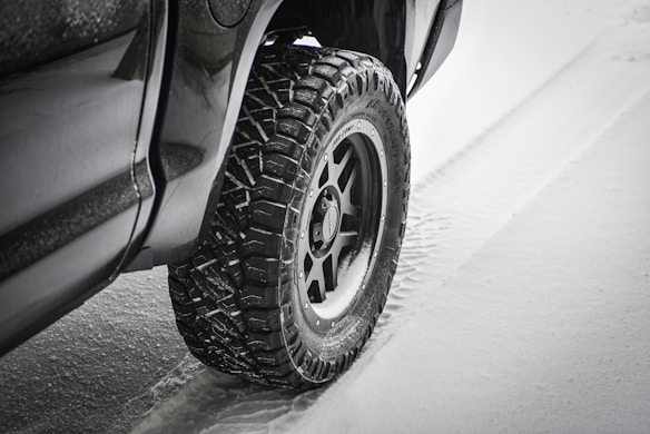 A close-up of a vehicle's rugged tire on a snowy surface, creating tracks in the snow. The tire features deep treads suited for off-road or winter conditions.