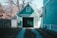 A small, green garage is nestled between two fences and surrounded by bare trees. The structure features vertical siding, a large dark door, and a triangular roof with a small window. Sunlight casts distinct shadows on the building, creating a tranquil suburban scene.