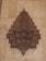 An intricately designed wooden ornament with geometric patterns and detailed perforations, mounted against a textured beige wall. The carved wooden structure features layers of symmetric, repeating shapes resembling a star or flower with multiple tiers.