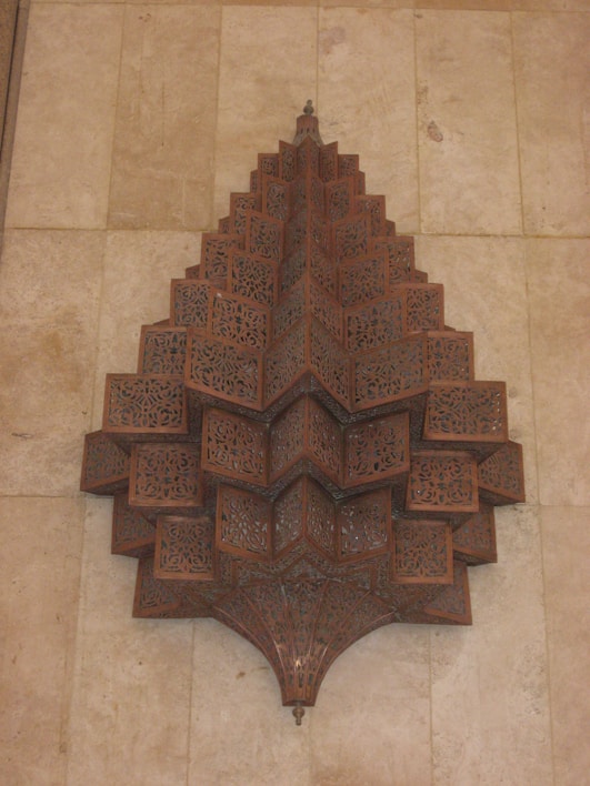 An intricately designed wooden ornament with geometric patterns and detailed perforations, mounted against a textured beige wall. The carved wooden structure features layers of symmetric, repeating shapes resembling a star or flower with multiple tiers.