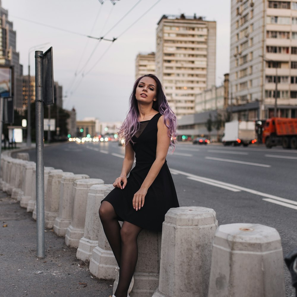 woman in black dress sitting on gray concrete post during daytime
