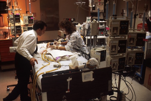 A medical setting with two people, likely healthcare professionals, focusing on a patient who is surrounded by numerous medical devices and monitors. The environment is busy with various tubes and equipment connected to the patient.