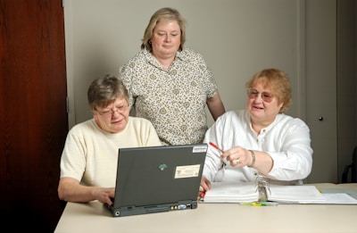 Three people are engaged in a collaborative setting, gathered around a laptop. Two are seated at a table, looking at the screen, while another stands nearby, observing. Papers and highlighters are spread on the table, indicating a work or learning environment.