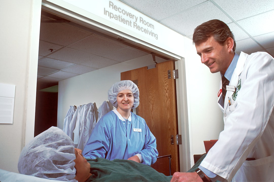 A Caucasian woman on a gurney just outside the surgery recovery room. A Caucasian physician and a nurse are on either side of the stretcher, either talking with her or pushing the gurney. The photograph was taken at lower than eye level.
