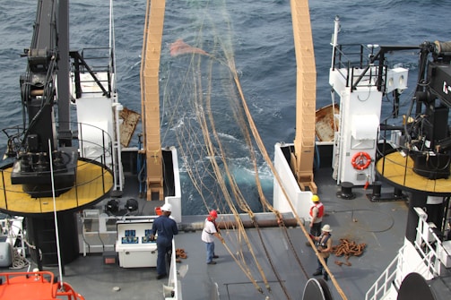 A group of people on a fishing boat deck, engaged in working with large fishing nets. The deck is equipped with cranes and various equipment. The ocean is visible in the background, indicating the boat is at sea.