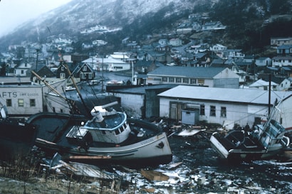 A coastal town is depicted in the aftermath of a disaster, with boats stranded on land amidst debris. The background shows a hillside with residential buildings. A notable presence of damaged structures and scattered wreckage suggests the severity of the event.