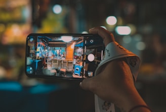 A hand holds a smartphone with the screen showing a live camera view of a small, colorful street food stall. The stall has bright lights, menus on the walls, and chairs in front. The background is blurred, indicating a nighttime setting, perhaps at a market or lively street.