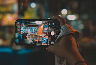 A hand holds a smartphone with the screen showing a live camera view of a small, colorful street food stall. The stall has bright lights, menus on the walls, and chairs in front. The background is blurred, indicating a nighttime setting, perhaps at a market or lively street.