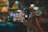A hand holds a smartphone with the screen showing a live camera view of a small, colorful street food stall. The stall has bright lights, menus on the walls, and chairs in front. The background is blurred, indicating a nighttime setting, perhaps at a market or lively street.