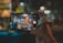 A hand holds a smartphone with the screen showing a live camera view of a small, colorful street food stall. The stall has bright lights, menus on the walls, and chairs in front. The background is blurred, indicating a nighttime setting, perhaps at a market or lively street.