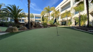 A well-maintained putting green is surrounded by lush palm trees and landscaped rocks, set against the backdrop of a modern building with large balconies. The sky is clear with a few wispy clouds.