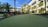 A well-maintained putting green is surrounded by lush palm trees and landscaped rocks, set against the backdrop of a modern building with large balconies. The sky is clear with a few wispy clouds.