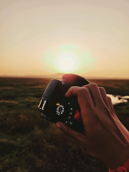 A hand holding a camera against a backdrop of a serene sunset over a grassy landscape. The sky is clear, and the sun is near the horizon, casting warm hues over the scene. The camera's settings dial is clearly visible.