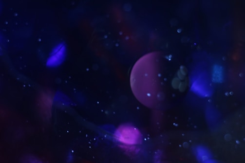 A dreamy and abstract scene featuring a dark background filled with various shades of blue and purple. A prominent spherical shape, resembling a planet, is visible in the center, surrounded by specks of light that appear like stars or cosmic dust.