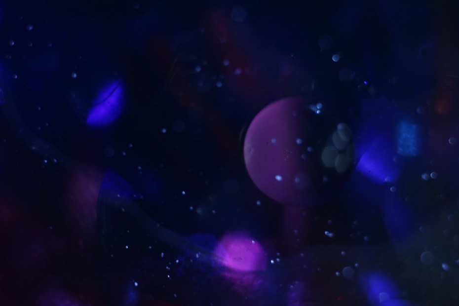 A dreamy and abstract scene featuring a dark background filled with various shades of blue and purple. A prominent spherical shape, resembling a planet, is visible in the center, surrounded by specks of light that appear like stars or cosmic dust.
