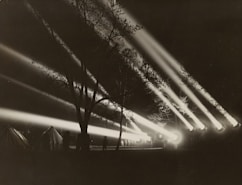 Strong beams of light pierce through the night sky, illuminating a dark forested area. In the foreground, several tents are visible along with blurred silhouettes of people. The scene suggests a heightened sense of urgency or alertness, possibly an outdoor event or military exercise.