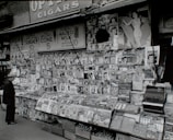 A vintage newsstand is packed with rows of magazines and newspapers, featuring vibrant covers with people and advertisements. A man wearing a hat stands in front, appearing to browse the selection. Above the stand, signs advertise soda, sundaes, ice cream cones, and malts, with prices clearly displayed. A Coca-Cola sign is partially visible in the upper corner.