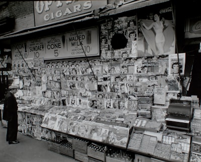 A vintage newsstand is packed with rows of magazines and newspapers, featuring vibrant covers with people and advertisements. A man wearing a hat stands in front, appearing to browse the selection. Above the stand, signs advertise soda, sundaes, ice cream cones, and malts, with prices clearly displayed. A Coca-Cola sign is partially visible in the upper corner.
