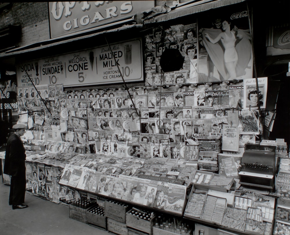 grayscale photo of man standing beside wall of magazines and newspapers
