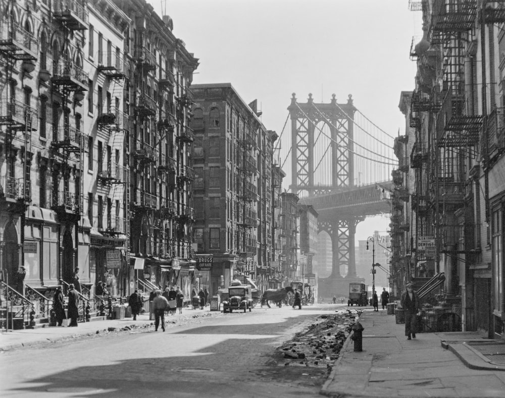 grayscale photo of people walking on street near buildings and bridge
