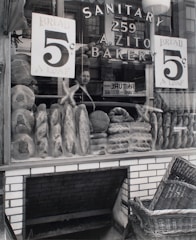 A bakery storefront displays an array of loaves and bread with signs advertising the price of 5 cents per loaf. The glass window shows reflections of a person and surrounding buildings. The store name 'A. Zito Bakery' is prominently displayed along with the word 'Sanitary'. Below the window, a woven basket rests near a brick base, suggesting a traditional or vintage shop setting.