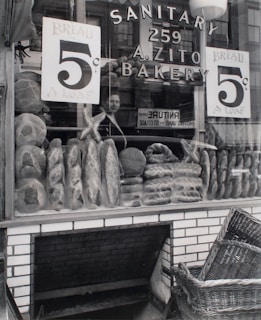 A bakery storefront displays an array of loaves and bread with signs advertising the price of 5 cents per loaf. The glass window shows reflections of a person and surrounding buildings. The store name 'A. Zito Bakery' is prominently displayed along with the word 'Sanitary'. Below the window, a woven basket rests near a brick base, suggesting a traditional or vintage shop setting.