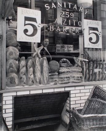 A bakery storefront displays an array of loaves and bread with signs advertising the price of 5 cents per loaf. The glass window shows reflections of a person and surrounding buildings. The store name 'A. Zito Bakery' is prominently displayed along with the word 'Sanitary'. Below the window, a woven basket rests near a brick base, suggesting a traditional or vintage shop setting.