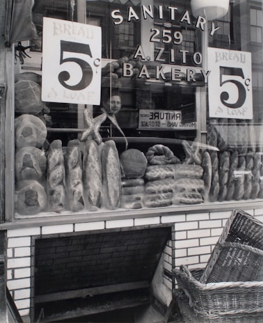 A bakery storefront displays an array of loaves and bread with signs advertising the price of 5 cents per loaf. The glass window shows reflections of a person and surrounding buildings. The store name 'A. Zito Bakery' is prominently displayed along with the word 'Sanitary'. Below the window, a woven basket rests near a brick base, suggesting a traditional or vintage shop setting.