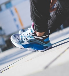 A close-up view of a person wearing stylish sneakers with a modern design, featuring a mix of blue, white, and black colors. The person is crouching, and the background is slightly blurred, suggesting an urban setting.