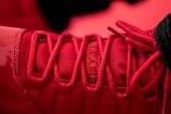 A close-up of a red sneaker, focusing on the detailed lacing and texture of the fabric. The shoe's branding is visible in black writing on the tongue.