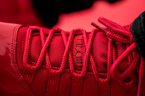 A close-up of a red sneaker, focusing on the detailed lacing and texture of the fabric. The shoe's branding is visible in black writing on the tongue.