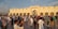 A large gathering of people outside a grand mosque with ornate architectural features and arched doorways. The diverse crowd is dressed in varying styles, with many wearing traditional attire. The sky is partly cloudy, and the sunlight casts long shadows on the polished surface of the plaza.