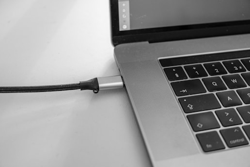 A close-up view of a laptop computer showing part of the keyboard and a connected charging cable. The keyboard features black keys with white symbols, and the charging cable is inserted into the side of the laptop.