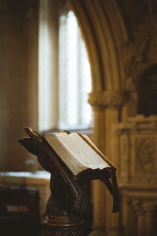 An ornate wooden lectern holds an open, aged book inside a dimly-lit, classical architectural setting with decorative arches and columns.