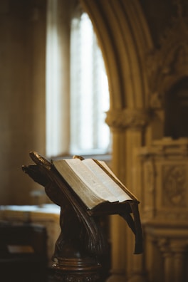 An ornate wooden lectern holds an open, aged book inside a dimly-lit, classical architectural setting with decorative arches and columns.