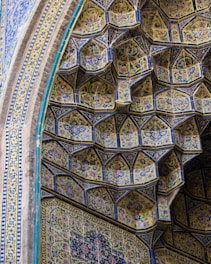 The image depicts a section of an ornate architectural structure, featuring intricate tile work in geometric patterns. The tiles, predominantly in shades of blue and yellow, are embellished with floral designs and arranged in a tiered, honeycomb-like formation. The craftsmanship demonstrates a blend of repetitive floral and arabesque motifs that create a harmonious and aesthetically pleasing visual effect.
