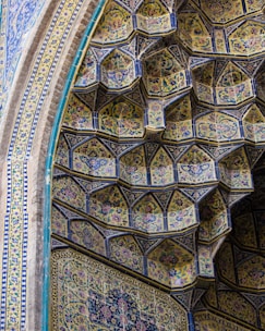 The image depicts a section of an ornate architectural structure, featuring intricate tile work in geometric patterns. The tiles, predominantly in shades of blue and yellow, are embellished with floral designs and arranged in a tiered, honeycomb-like formation. The craftsmanship demonstrates a blend of repetitive floral and arabesque motifs that create a harmonious and aesthetically pleasing visual effect.