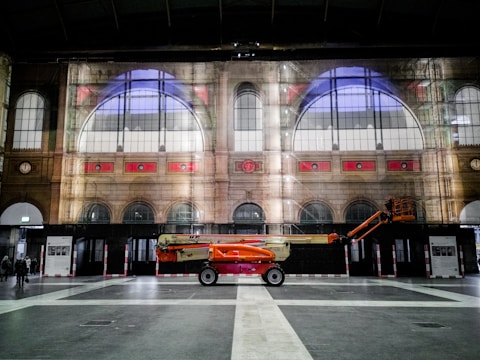 An indoor scene of a grand architectural structure with large arched windows and intricate interior design. A bright orange lift vehicle is parked on the polished floor, creating a stark contrast with the muted colors of the building. People are seen walking at a distance, adding a sense of scale to the space.