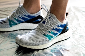 A pair of modern athletic shoes featuring a textured gray mesh upper with accents of blue and teal stripes. The shoes have a white cushioned sole and are being worn by a person standing on a metallic, reflective surface. The laces are tightly tied, ensuring a secure fit.