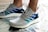 A pair of modern athletic shoes featuring a textured gray mesh upper with accents of blue and teal stripes. The shoes have a white cushioned sole and are being worn by a person standing on a metallic, reflective surface. The laces are tightly tied, ensuring a secure fit.