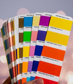 A hand holds a fanned-out selection of Pantone color swatches. The swatches include a variety of vibrant colors, such as pink, yellow, green, blue, orange, and red. The background is softly blurred, enhancing the focus on the colorful swatches.