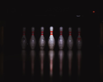 A row of seven bowling pins is aligned on a reflective surface, illuminated against a dark background. The pins have red stripes and show signs of wear and use.